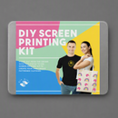 A mockup of a DIY Screen Printing Kit box shown on a neutral gray background. The packaging features a colorful geometric design in pink, green, yellow, and blue, with a photo of a man and woman holding a printed shirt.